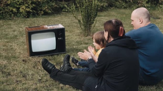 一家人坐在草地上，看着古老的复古电视。他们拍手