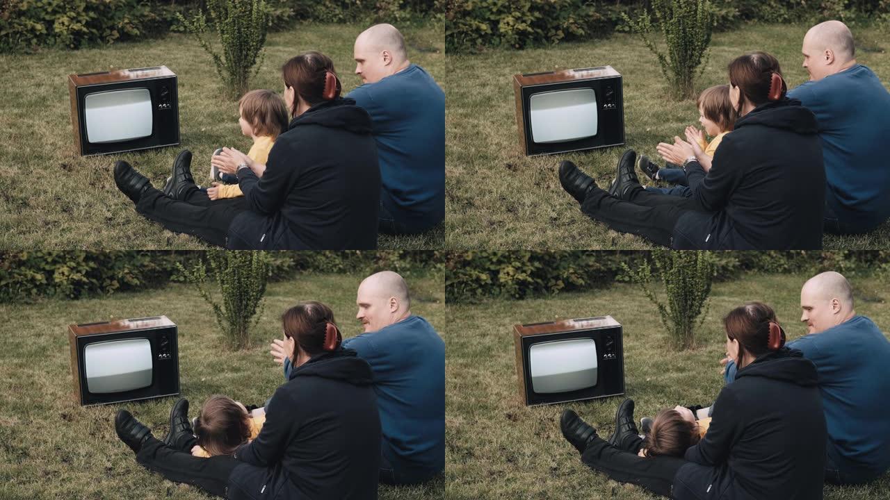 一家人坐在草地上，看着古老的复古电视。他们拍手