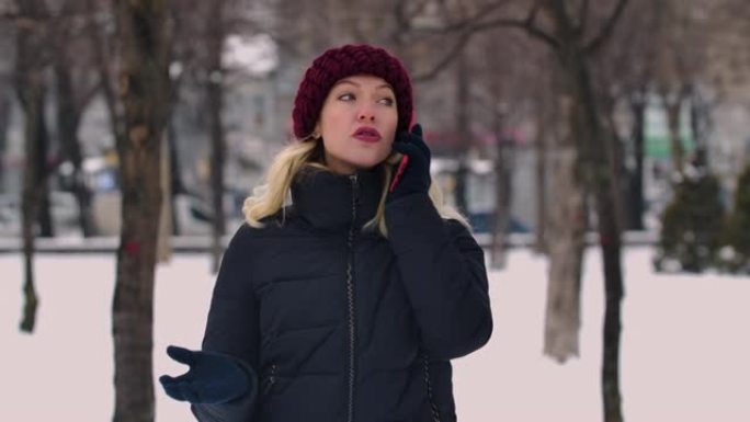 一个年轻迷人的金发女子正在用手机聊天，环顾四周。一个女孩站在白雪皑皑的街道中间。背景模糊了。慢动作。
