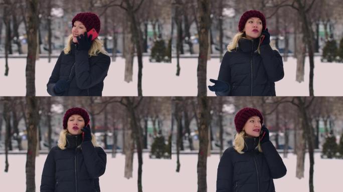 一个年轻迷人的金发女子正在用手机聊天，环顾四周。一个女孩站在白雪皑皑的街道中间。背景模糊了。慢动作。