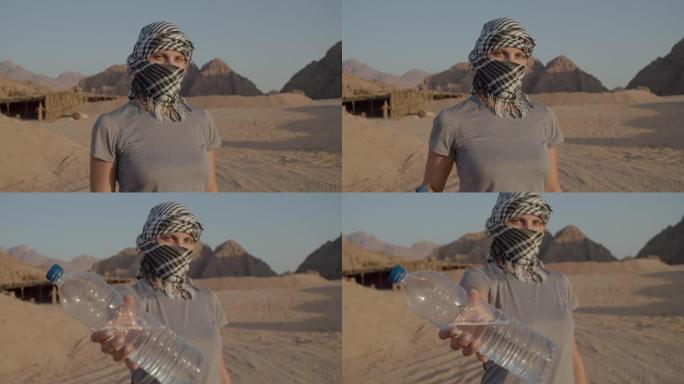 穿着方格keffiyeh的女人拿着一瓶站在沙漠中的水。带塑料水瓶的高加索女游客。饮用水的重要性。
