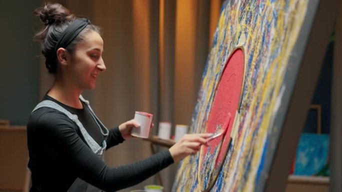 微笑的女人站在画架附近，画布上正在创作最新的展览绘画。女人努力完成杰作，用附加的圆形模板的红色油漆填