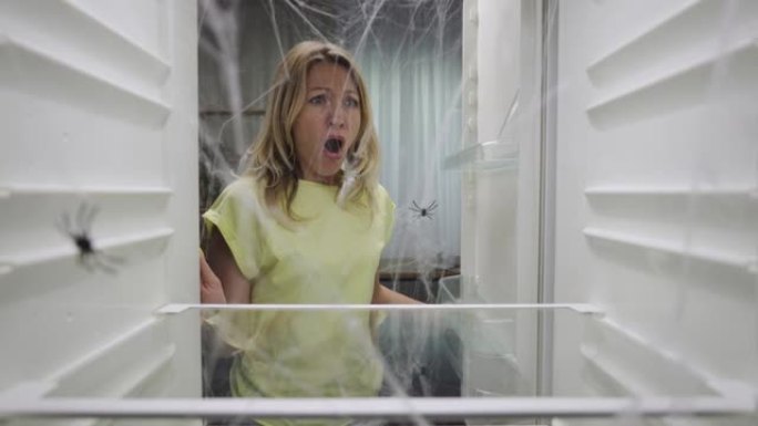 女人打开冰箱门，看到蜘蛛网里面有蜘蛛而不是食物，然后害怕地关上了。一个空的、可怕的冰箱的内部视图。特