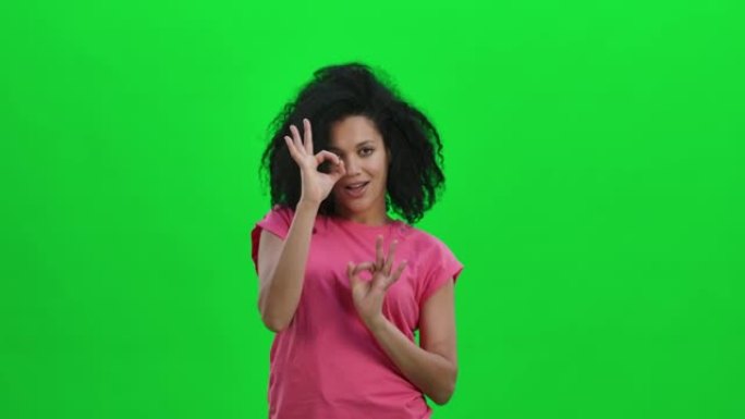 年轻女性非洲裔美国人微笑并做出标志的肖像。卷发的黑人妇女在工作室的绿色屏幕上摆姿势。特写。慢动作就绪