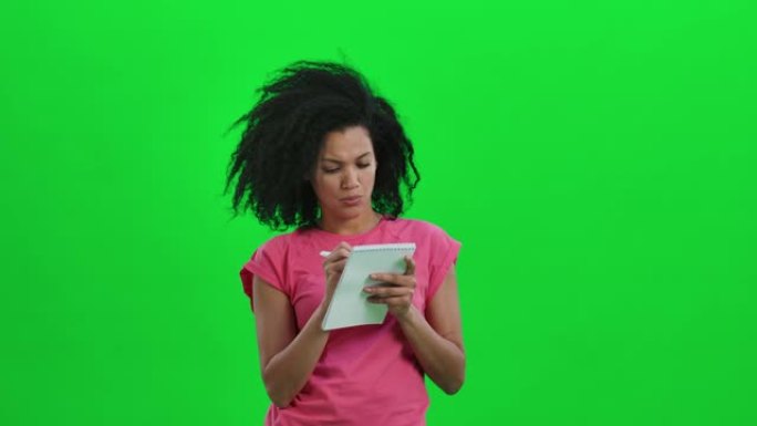年轻女性非裔美国人思考的肖像，然后在笔记本上用笔愉快地书写。卷发的黑人妇女在工作室的绿色屏幕上摆姿势