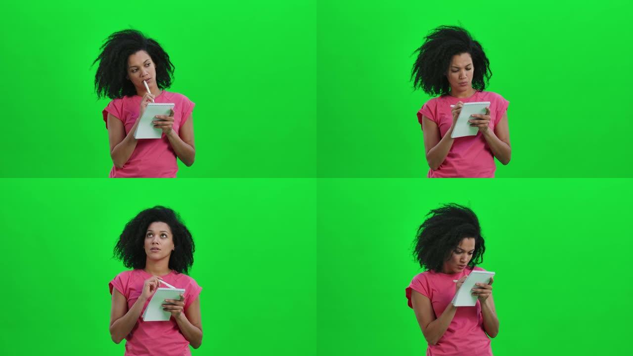 年轻女性非裔美国人思考的肖像，然后在笔记本上用笔愉快地书写。卷发的黑人妇女在工作室的绿色屏幕上摆姿势