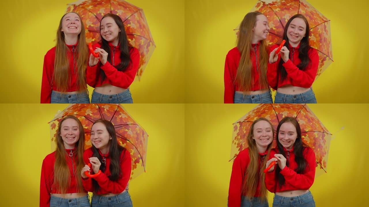 美丽的双胞胎姐妹笑着看着黄色背景下的相机旋转伞。轻松快乐的白人女性玩得开心。个性与生活理念