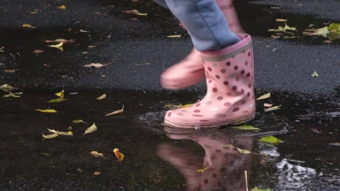 穿着胶靴的女童或青少年在覆盖着水坑和落叶的湿沥青上奔跑。儿童和秋天。全高清慢动作视频