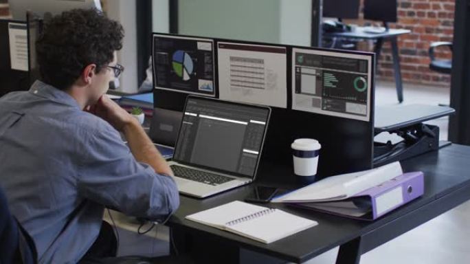 白人男子坐在办公桌前观看笔记本电脑和计算机屏幕上的编码数据处理