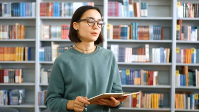 一位年轻迷人的女老师授课的特写肖像，在大学图书馆书架背景上的笔记本上阅读作业