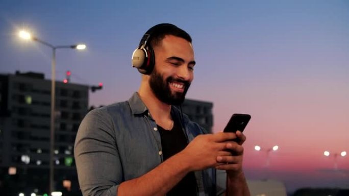 年轻的大胡子跨国男人一边戴着耳机听音乐一边放松。在美丽的日落中行走，微笑着使用智能手机。社交网络。浏