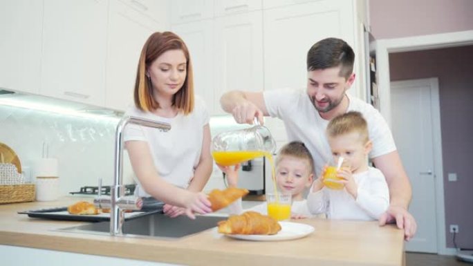 年轻幸福的家庭父母和两个开朗的小儿子在家里的厨房里一起喝热饮。