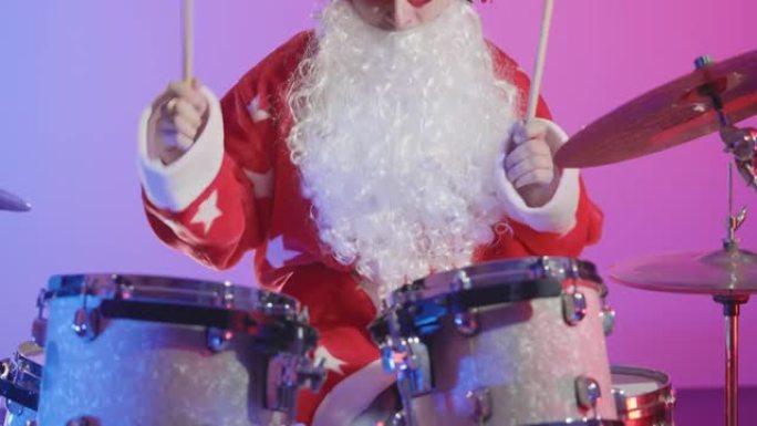 一个打扮成圣诞老人的男人在黑暗的工作室里在明亮的紫色灯光背景下演奏鼓包。穿着狂欢节服装的音乐家在圣诞