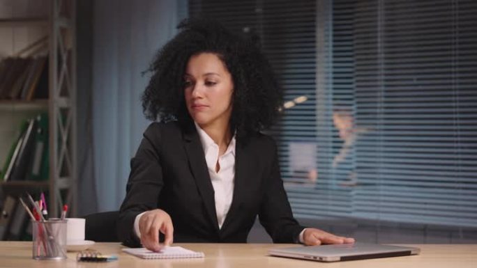 非裔美国妇女的肖像完成工作，关闭笔记本电脑并离开。女商人在工作场所摆姿势，坐在办公室内部的桌子旁。慢