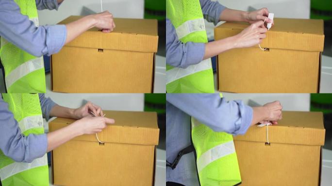 穿着安全衬衫的女人的手正在使用白色塑料封条来锁定棕色纸盒，以防止文件盒被非法打开。