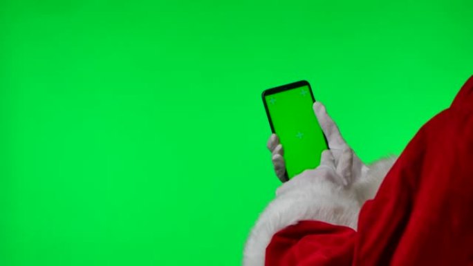 圣诞老人用智能手机戴上白手套，轻扫并点击绿屏色度键。孤立在工作室的绿色背景上。模拟显示。特写。慢动作