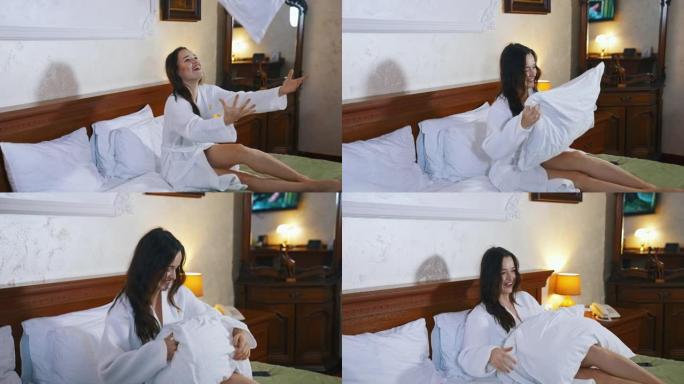 酒店床上的美女。穿着白色浴袍的快乐年轻女子坐在床上，扔枕头。愉快的假期。
