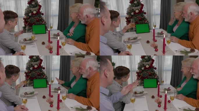 幸福的家人或朋友坐在圣诞节装饰的桌子旁，用酒杯敬酒，并在节日期间使用智能手机进行视频通话。绿屏