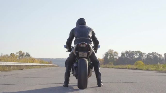 戴着防护头盔的年轻骑自行车的人的后视图正坐在乡间小路的摩托车上，准备骑行。摩托车装备的摩托车手将驾驶