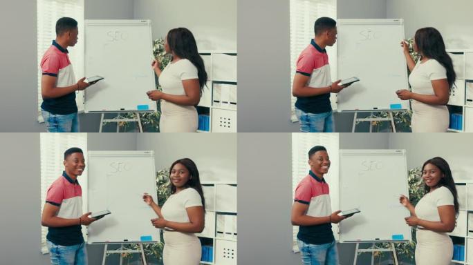 两个朋友，学生，皮肤黝黑的同事站在白板旁边，女孩拿着记号笔画画，男孩检查平板电脑上的图形，他们看着相
