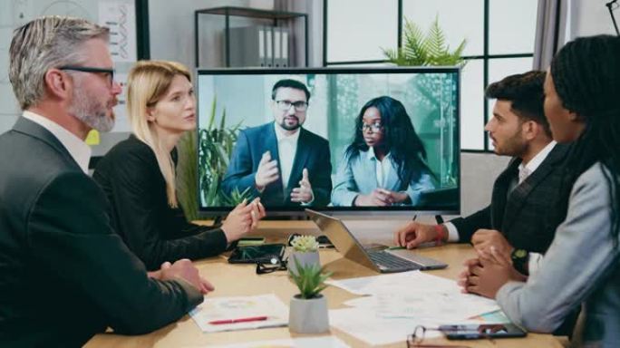 好看的聪明的专业经验丰富的混合种族商人在电视上与男女国际合作伙伴在办公室的视频会议期间为自己提供建议