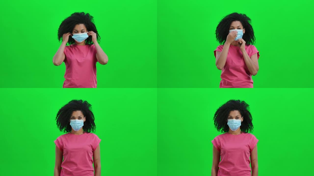 年轻女性非洲裔美国人的肖像看着相机，戴上医用口罩。卷发的黑人妇女在工作室的绿色屏幕上摆姿势。特写。慢