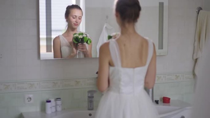 浪漫快乐苗条的年轻新娘在浴室镜子里反射，闻到花束的微笑。优雅美丽的高加索女人仰慕早晨在室内的倒影。慢