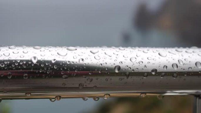 在模糊的海洋背景下，不锈钢扶手上的雨滴。特写镜头。雨露中的水滴聚集在闪亮的钢铬杆手轨栏杆上