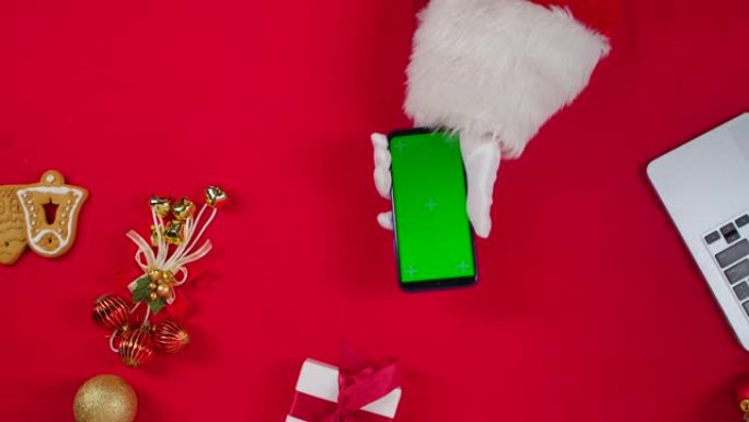 顶视图圣诞老人的手使用智能手机，并在红色新年装饰桌旁喝茶。圣诞老人点击绿屏色度键观看内容，进行在线购