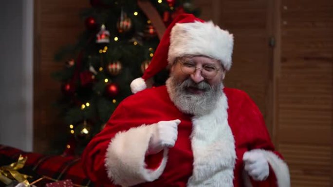 圣诞老人跳舞滑稽的肖像。老人留着红色西装，戴着帽子，在圣诞树的背景下戴着玩具和灯。新年快乐的夜晚。慢