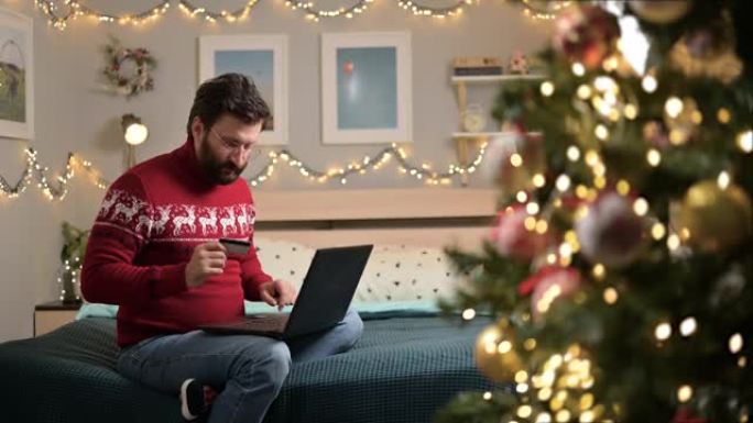圣诞礼物。网上购买商品。圣诞节前夕，一名男子坐在家里的床上，拿着笔记本电脑用信用卡付款。他对高昂的价