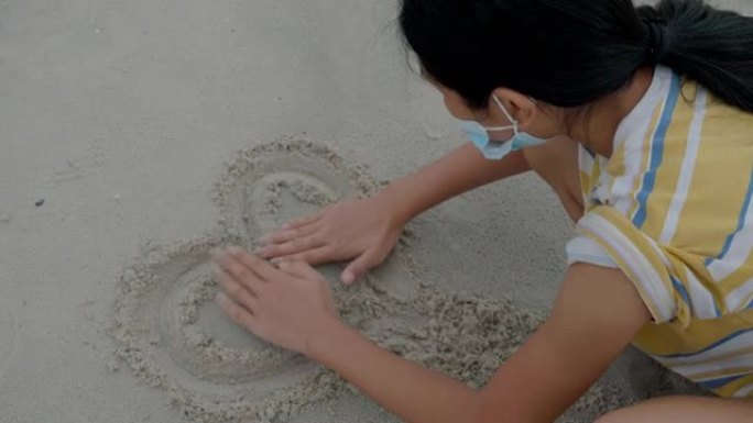 亚洲女孩戴着口罩，在沙滩上画心形，新的正常生活方式理念。