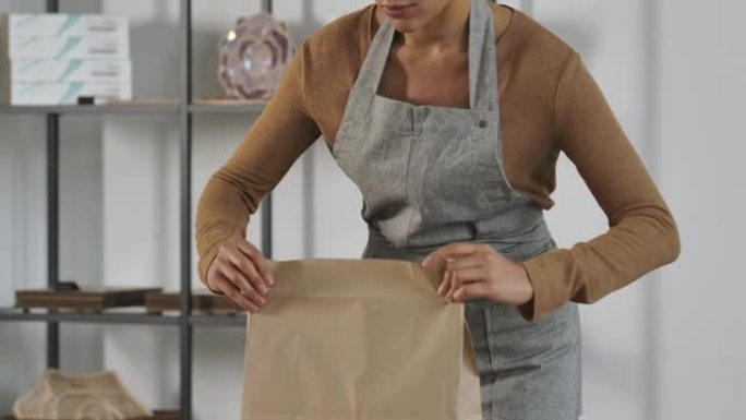 年轻女孩在商店里用网上订单拿起一个纸袋。一位可爱的非洲裔美国妇女在灯光室的背景下为顾客包装商品。特写
