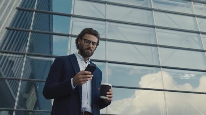 年轻的跨国男子戴着眼镜在智能手机上聊天。输入消息。走过大工业大楼。喝咖啡去。真诚地微笑。古典夹克。应
