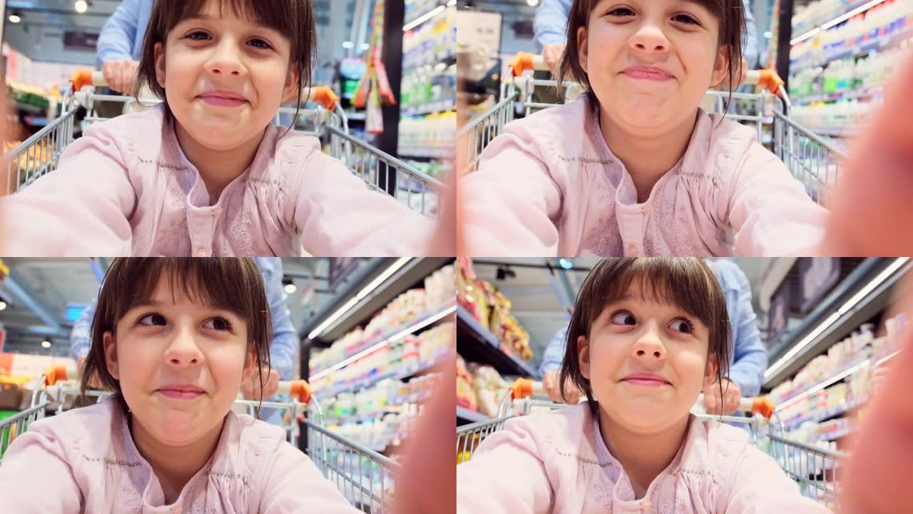 快乐的女孩拿着相机，坐在杂货店里拍摄自己，她的母亲在商店里推着相机。