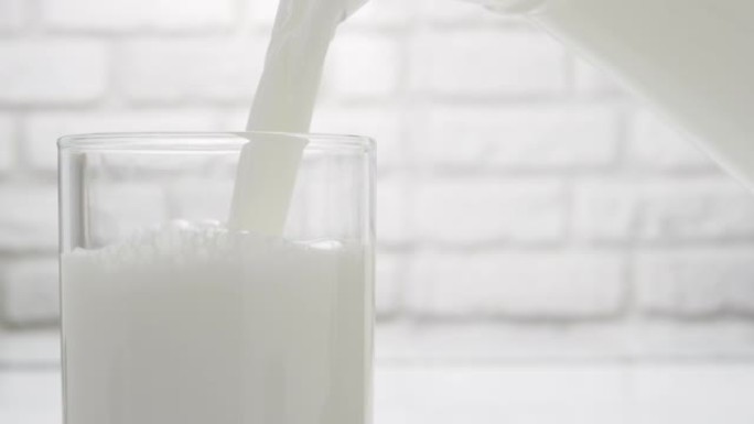 在白色砖墙的背景下，从水罐中倒入牛奶。乳制品特写。牛奶流入玻璃杯