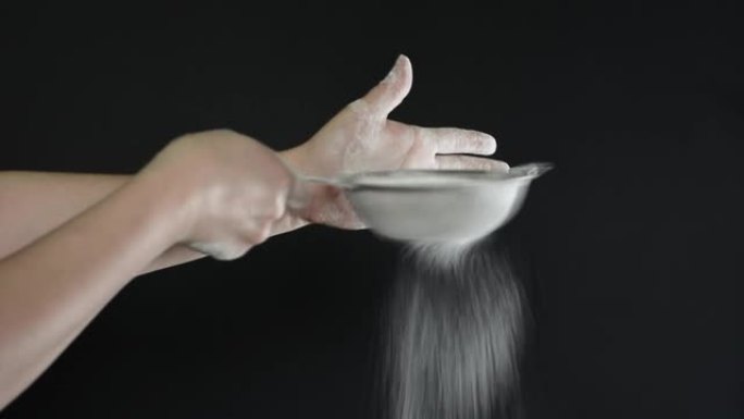 厨房里的女人通过黑色背景特写上的金属筛筛选类似于面粉或糖的白色粉末