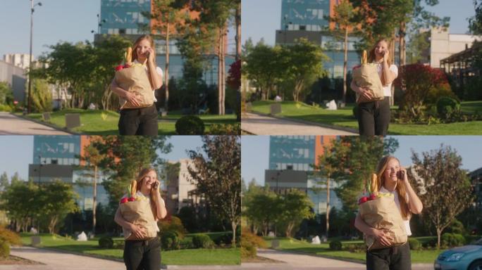 在镜头前走在街上的金发女人拿着装满蔬菜和水果的生态袋，她在电话里说话，享受阳光。4k