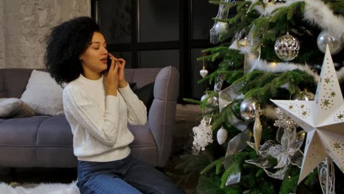 一个快乐的非洲裔美国妇女在智能手机上聊天的肖像。年轻女子坐在明亮的阁楼风格房间里的节日圣诞树旁。节日