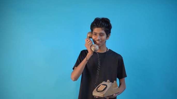 穿着黑色t恤的英俊剃光的印度年轻人20多岁，在工作室里用老式电话孤立在蓝色背景上。