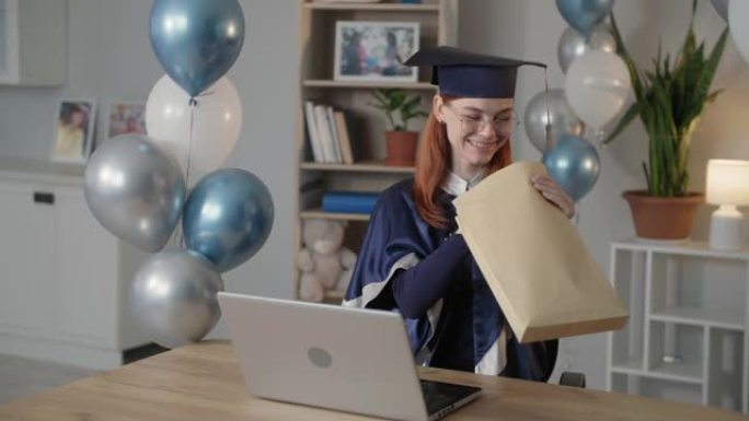 在线毕业，穿着学术服装和帽子的快乐年轻女子通过笔记本电脑上的视频通话进行交流，并在大流行期间向家人展