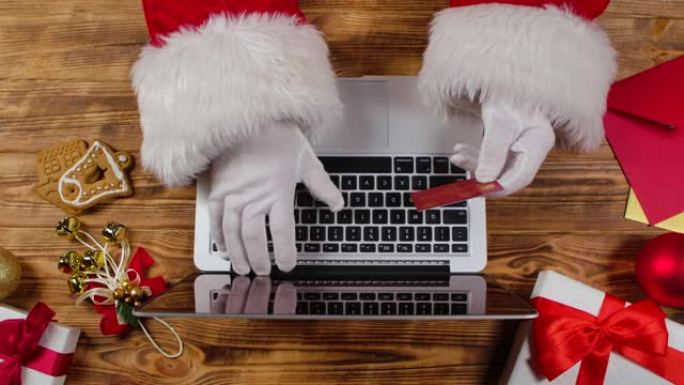 顶视图圣诞老人戴着白手套的手在木制新年装饰桌子上的键盘上打字。圣诞老人与笔记本电脑一起工作，同时使用