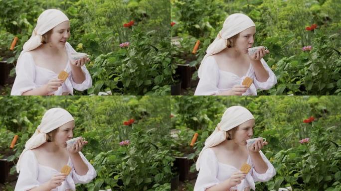 暑假，穿着头巾的乡村少女在乡村花园户外喝杯饼干茶。美丽的农村妇女在夏日村喝茶，拿着饼干。