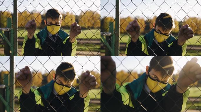 一个戴着医用口罩的男孩用手抓住了链环网，并用手摇动了篱笆。他在篱笆的另一边