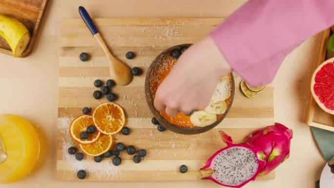 女人用手将种子撒在水果粥的特写镜头上，用浆果和柑橘制成的橙色冰沙碗的俯视图。健康早餐。不含糖的甜食