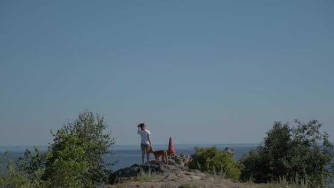 一名年轻女子带着法老品种的红狗沿着海岸旅行。夏天阳光明媚的日子，蓝天。