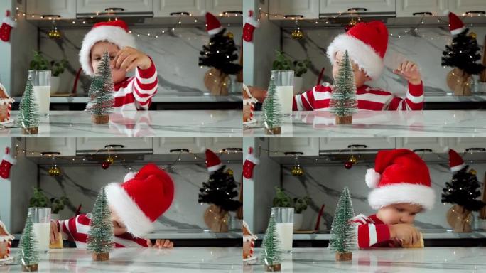 戴着圣诞老人帽子的有趣的蹒跚学步的男孩在厨房里喝牛奶，吃饼干，装饰圣诞节。假期、传统、婴儿食品和健康