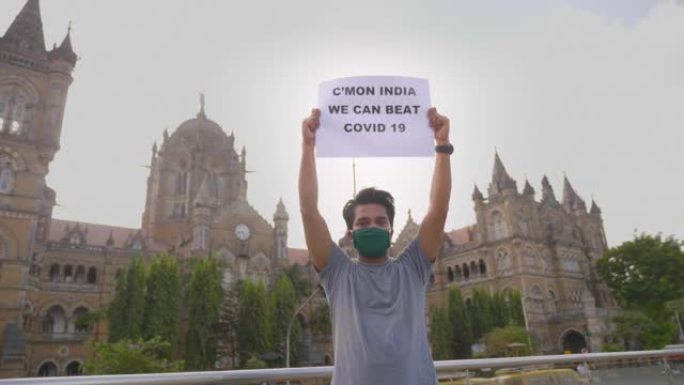 一个戴着口罩的年轻人站着，举着标语牌，上面写着 “来吧，印度，我们可以击败Covid 19”。