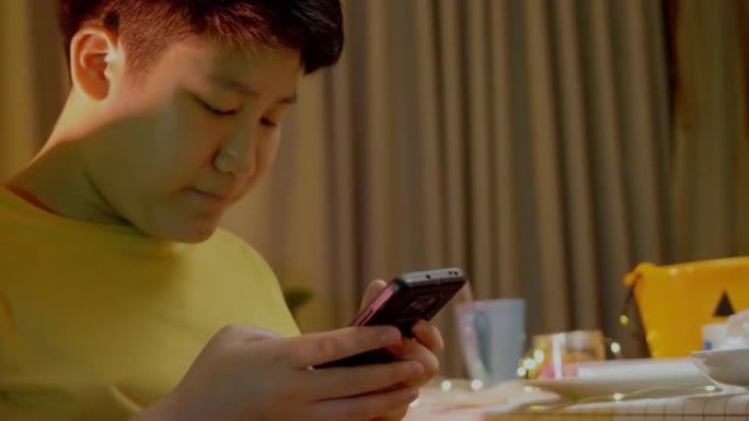 亚洲男孩在晚上在家玩手机游戏，带圣诞灯背景。