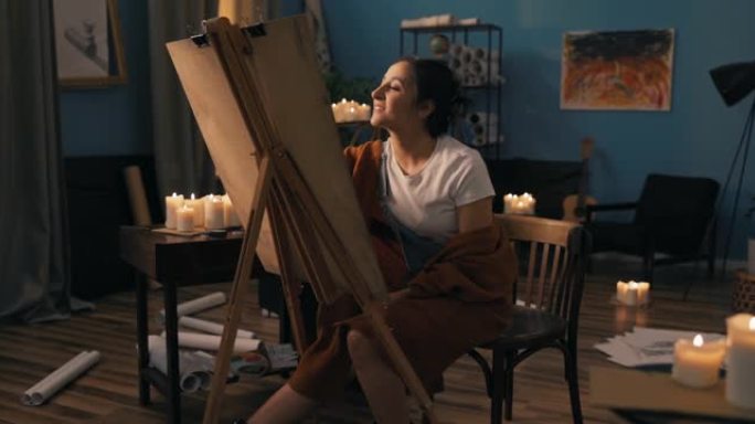 艺术系学生坐在远处教室的家中为大学画项目美丽的女人拿着画布站在画架上分析完成的草图最后的触摸微笑看着
